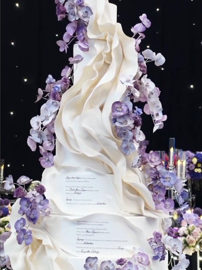 Kue pengantin pun diberi sentuhan personal. LeNovelle Cake mengkreasikan wedding cake putih berhias bunga anggrek dengan penggalan lirik lagu duet mereka. Foto: Instagram @thebridestory @johan.k.lotus