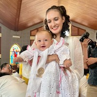 Walaupun masih tampak kebesaran namun baby Djala tampak imut dan menggemaskan mengenakan dress putih tersebut. Sambil digendong sang mama, baby Djala tampak tersenyum. Foto: dok. Instagram/@nadinelist