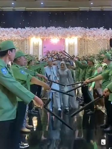 Acara pernikahan ini mendadak viral di media sosial karena pasangan pengantin ini berjalan di tengah-tengah barisan para pria yang memakai topi serta seragam seperti hansip