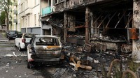 Beberapa mobil yang diparkir di seberang jalan dari gedung yang sebagian hancur terbakar dan puing-puing terlihat berserakan di area yang luas.