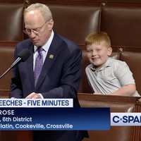 Bocah yang baru berusia 7 tahun itu duduk di belakang ayahnya. Baru John Rose mulai berpidato di forum formal itu, anaknya langsung menyadari kamera juga sedang menyorot ke arah mereka. (Foto: YouTube/C-Span)