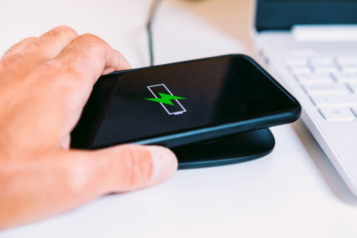 Hand with black mobile smart phone with the logo of a battery with green lightning on the screen, charging on a wireless charger base next to a laptop on a white work table