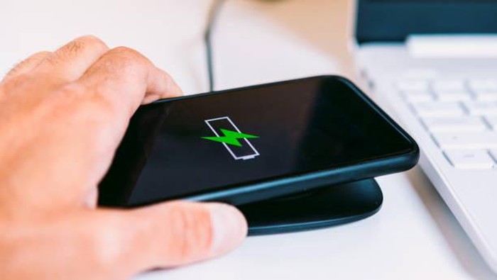 Hand with black mobile smart phone with the logo of a battery with green lightning on the screen, charging on a wireless charger base next to a laptop on a white work table
