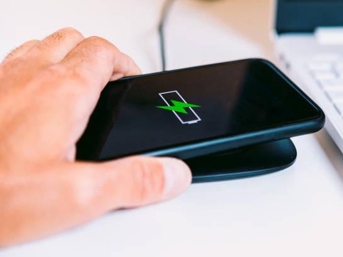 Hand with black mobile smart phone with the logo of a battery with green lightning on the screen, charging on a wireless charger base next to a laptop on a white work table