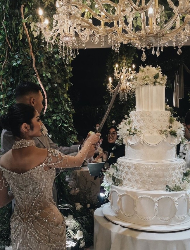 Untuk kue pengantinnya Marco dan Debi memilih kue berwarna putih dengan 5 tingkat. Sesuai dengan tema resepsi pernikahan, kue tersebut juga dihiasi dengan bunga-bunga artificial berwarna senada. Foto: Instagram
