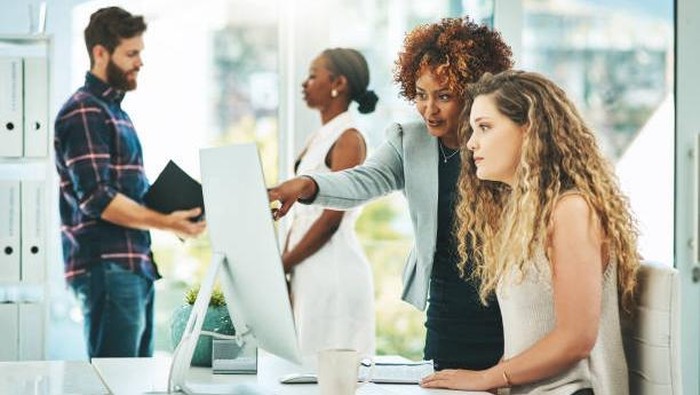 Shot of two businesswomen working together on a computer in an office with their colleagues in the background
