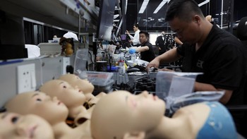 Masker silikon sepanjang leher tergeletak di atas meja di samping lengan dan kaki silikon, sementara kepala tanpa tubuh dipajang dan robot humanoid dalam berbagai tahap konstruksi berdiri di dekatnya.