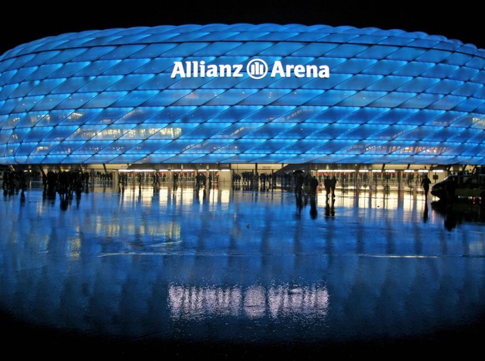 MUNICH, Germany:  The illuminated new Allianz Arena Football stadium in Munich is reflected in the wet road 30 May 2005 after a friendly football match between TSV 1860 Munich and FC Nuremberg, one day before the Arenas official opening. The Arena, with a capacity of 66.000 spectators, was built at a cost of EUR 275 million and will be one of the stadiums to host 2006 Football World Cup matches. AFP PHOTO DDP/TIMM SCHAMBERGER    GERMANY OUT    Bei stroemenden Regen verlassen am Montag (30.05.05) Fussballfans die Eroeffnungsveranstaltung der neuen Muenchner Allianz-Arena. Zur Eroeffnung fand ein Fussballspiel zwischen dem TSV 1860 Muenchen und dem 1. FC Nuernberg statt. Foto: Timm Schamberger/ ddp  (Photo credit should read TIMM SCHAMBERGER/DDP/AFP via Getty Images)
