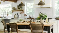 Dapur yang terang dan lapang ini memiliki kabinet putih, dinding belakang putih, dan kabinet serta hood cooker yang berwarna putih. Kursi dengan sandaran rotan dan meja hitam dari kayu menampilkan gaya pedesaan Prancis. Foto: JOYELLE WEST via Better Homes & Gardens