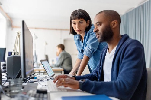 Two business people looking at desktop computer monitor and discussing new programme codes. Male professional working on computer with female colleague standing by looking at computer monitor.