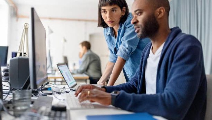 Two business people looking at desktop computer monitor and discussing new programme codes. Male professional working on computer with female colleague standing by looking at computer monitor.