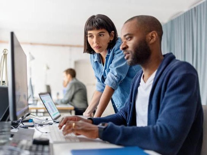 Two business people looking at desktop computer monitor and discussing new programme codes. Male professional working on computer with female colleague standing by looking at computer monitor.