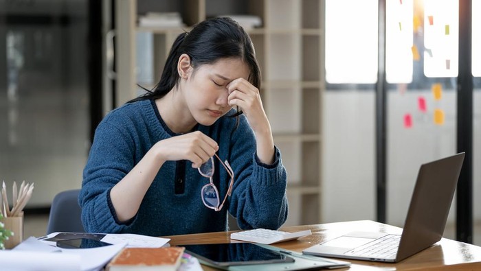 Frustrated young woman feeling tired and stressed, keeping eyes closed and massaging nose while sitting at her working place.
