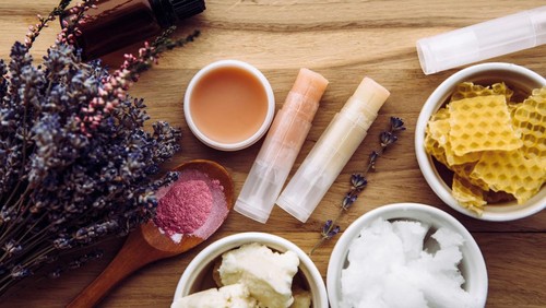 Ingredients for homemade lip balm: shea butter, essential oil, mineral color powder, beeswax, coconut oil. Homemade lip balm lipstick mixture with ingredients scattered around.