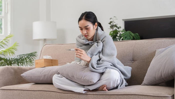 Young woman feeling unwell, wrapped in a blanket, checking her temperature with a thermometer while sitting on a couch at home.