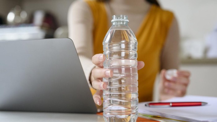 Surface level close-up of Caucasian female telecommuter reaching for uncapped bottle of water as she takes a break from using laptop.