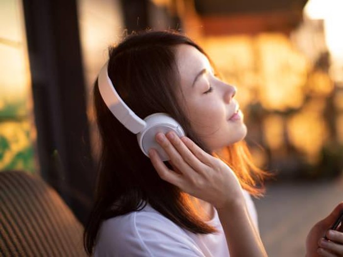 Portrait of smiling young Asian woman with eyes closed enjoying music over headphones and using smartphone, relaxing on the balcony against sunlight.