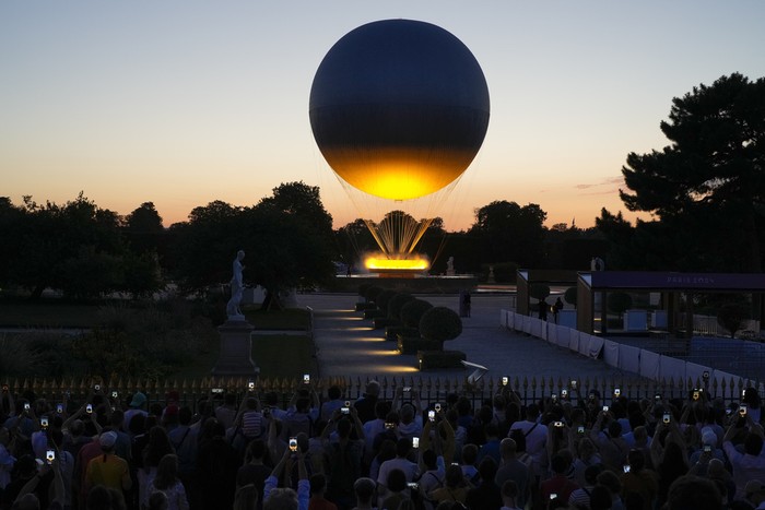 A crowd takes photos at the Tuileries Garden as the cauldron rises at sunset during the 2024 Summer Olympics, Sunday, July 28, 2024, in Paris, France. (AP Photo/Natacha Pisarenko)