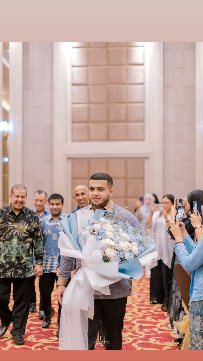 Yislam tampil mengenakan kemeja batik perpaduan warna biru keunguan dan coklat.  Kemeja lengan panjang itu dipadukan dengan celana panjang bahan berwarna hitam. Dalam foto yang dibagikan terlihat Yislam masuk ke ruangan sambil membawa sebuah buket bunga. Foto: Instagram