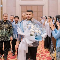 Yislam tampil mengenakan kemeja batik perpaduan warna biru keunguan dan coklat.  Kemeja lengan panjang itu dipadukan dengan celana panjang bahan berwarna hitam. Dalam foto yang dibagikan terlihat Yislam masuk ke ruangan sambil membawa sebuah buket bunga. Foto: Instagram