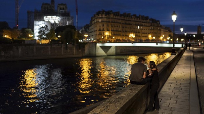 Victor, left, and his girlfriend, Florence, talk along the Seine River at dusk as the spire of the Notre Dame Cathedral rises in the background ahead of the 2024 Summer Olympics, Tuesday, July 16, 2024, in Paris, France. (AP Photo/David Goldman)
