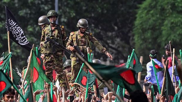 Para pengunjuk rasa antipemerintah berbaris menuju istana Perdana Menteri Sheikh Hasina sementara personel militer (tengah) berjaga di kawasan Shahbag, dekat Universitas Dhaka di Dhaka pada 5 Agustus 2024. Protes di Bangladesh yang dimulai sebagai demonstrasi yang dipimpin mahasiswa terhadap aturan perekrutan pemerintah pada bulan Juli mencapai puncaknya pada tanggal 5 Agustus, saat perdana menteri melarikan diri dan militer mengumumkan akan membentuk pemerintahan sementara. (Photo by Munir UZ ZAMAN / AFP)