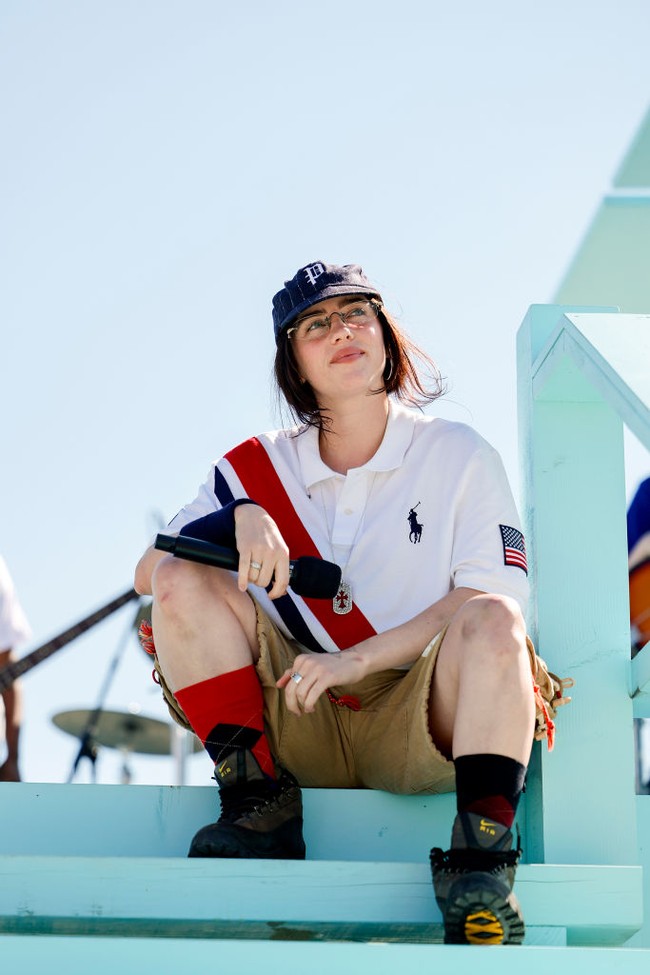 Billie Eilish memadukan kaus putih oversized bermotif garis diagonal biru-merah dengan celana pendek warna khaki. Topi baseball dan kacamata melengkapi penampilannya. Foto: Emma McIntyre/Getty Images for LA28