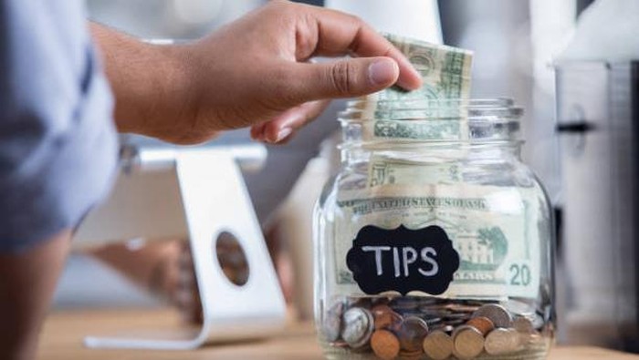 Unrecognizable male coffee shop customer places cash into a tip jar.