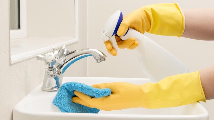 Hands in gloves spraying liquid soap on a rag for cleaning bathroom sink