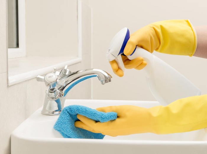 Hands in gloves spraying liquid soap on a rag for cleaning bathroom sink