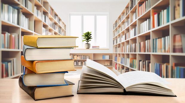 Book stack and opened book on the desk on blurred bookshelves in light public library room background