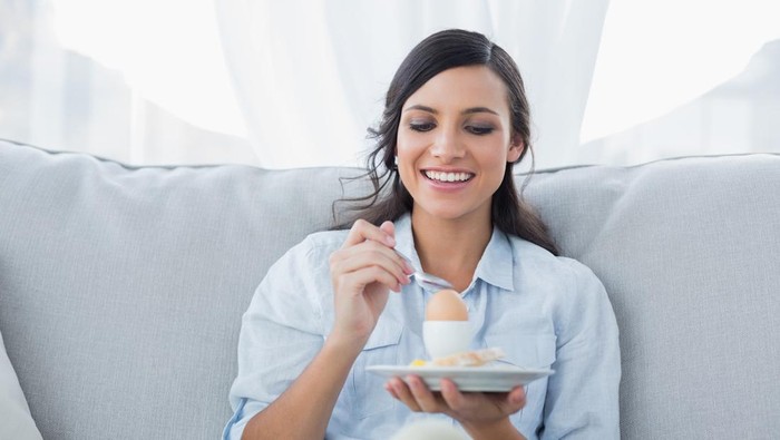 Cheerful brunette eating egg in her living room