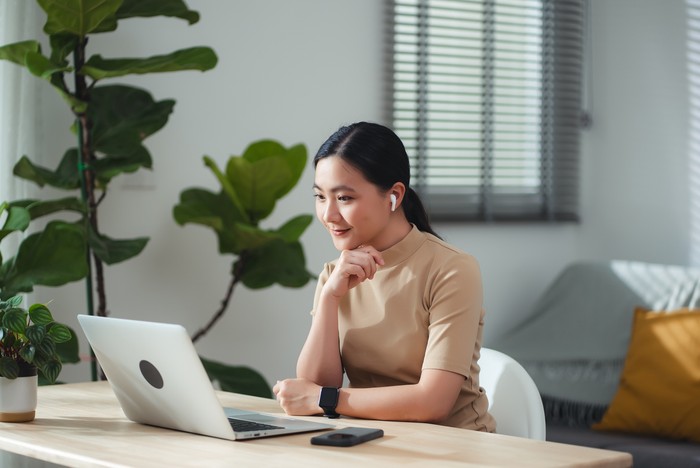 Asian woman in earbuds enjoy with work looking on laptop screen and thinking about project, sitting at home office. Happy woman working from home.