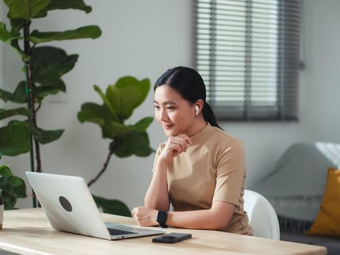 Asian woman in earbuds enjoy with work looking on laptop screen and thinking about project, sitting at home office. Happy woman working from home.