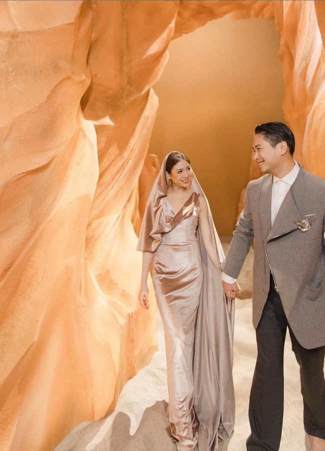 Studio dibuat seperti gurun pasir dengan palet warna camel. Clarissa tampak mengenakan gaun bertudung warna emas metalik rancangan desainer Stella Rissa. Sang suami, Anthony Tjipto, mengenakan jaket dengan lapel asimetris dipadu celana abu-abu gelap dan kemeja putih. Foto: Instagram/@clarissatanoe @solemnstudios