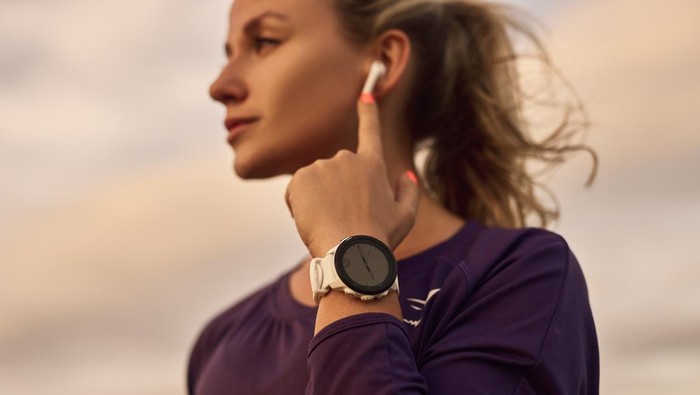 Low angle of young female in sportswear with smartwatch adjusting earbuds, while resting during outdoor fitness workout in summer evening with cloudy sky in background