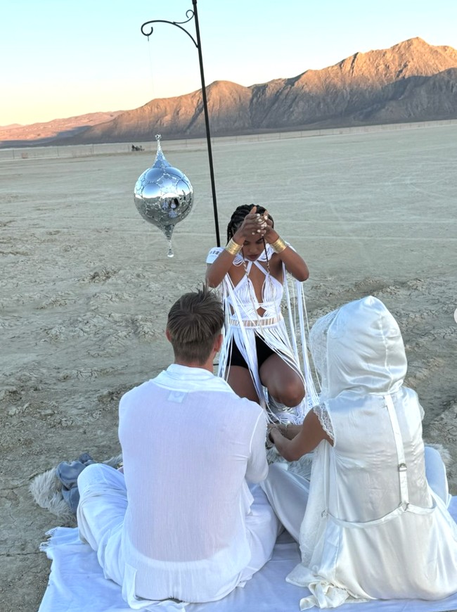 Dalam pernikahan mereka, memperlihatkan pasangan pengantin duduk di atas kain putih di tengah gurun, dengan seorang wanita mengenakan busana serba putih di hadapan mereka. Wanita ini tampak sedang melakukan gerakan tangan yang menyerupai ritual atau prosesi simbolis. Foto: dok. Instagram @kellygale