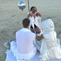 Dalam pernikahan mereka, memperlihatkan pasangan pengantin duduk di atas kain putih di tengah gurun, dengan seorang wanita mengenakan busana serba putih di hadapan mereka. Wanita ini tampak sedang melakukan gerakan tangan yang menyerupai ritual atau prosesi simbolis. Foto: dok. Instagram @kellygale