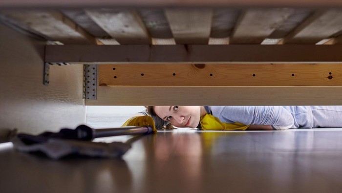 Woman in protective gloves cleaning floor under bed in room using flat wet mop. House cleaning.