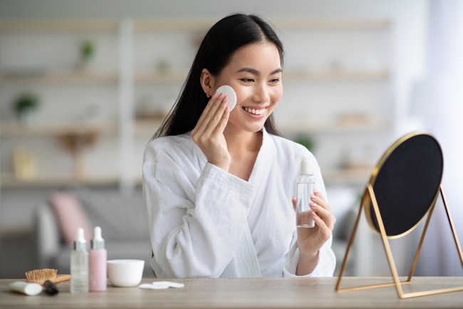 Cheerful asian woman cleaning her face, using cotton pads and cleansing product, looking at mirror in bedroom. Young attractive korean lady using face toner and cotton pad, home interior, empty space