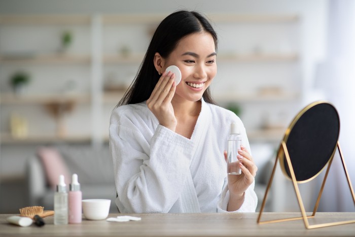 Cheerful asian woman cleaning her face, using cotton pads and cleansing product, looking at mirror in bedroom. Young attractive korean lady using face toner and cotton pad, home interior, empty space