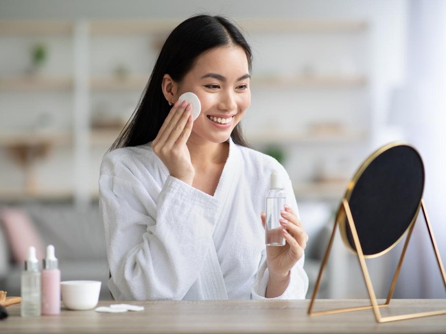 Cheerful asian woman cleaning her face, using cotton pads and cleansing product, looking at mirror in bedroom. Young attractive korean lady using face toner and cotton pad, home interior, empty space