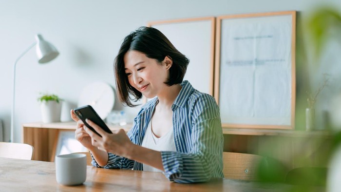 Beautiful smiling young Asian woman managing financial bill payment with smartphone and credit card in the living room at home. Technology makes life so much easier