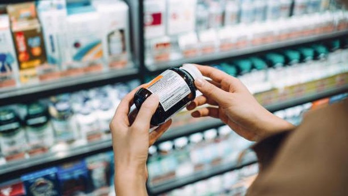 Over the shoulder view of young Asian woman browsing through medical products and reading the label on a bottle of medicine in front of the shelves in a pharmacy