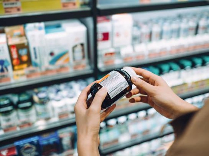 Over the shoulder view of young Asian woman browsing through medical products and reading the label on a bottle of medicine in front of the shelves in a pharmacy