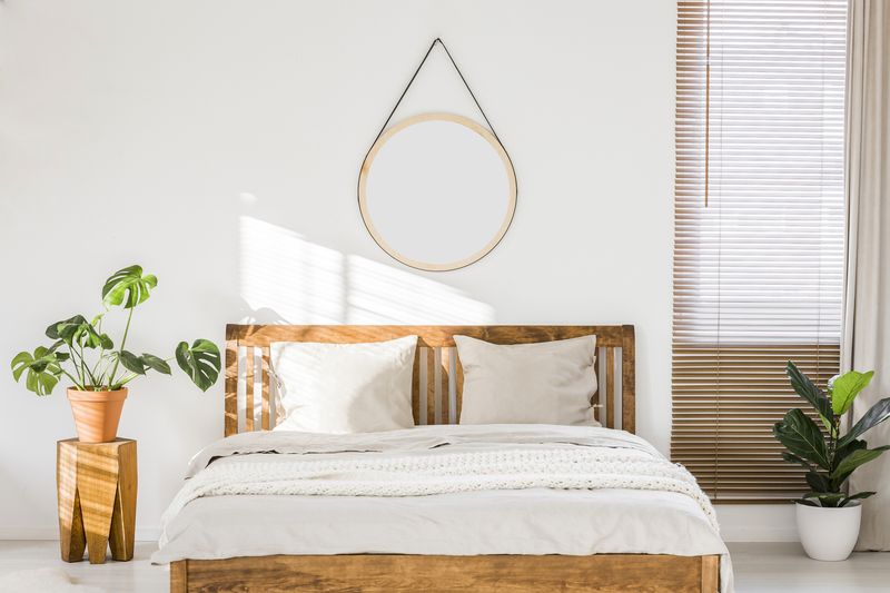 Sun shining on a white wall with a round mirror in a minimalist bedroom interior with natural, wooden furniture and beautiful green plants