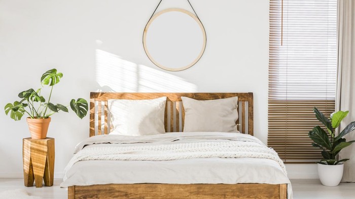 Sun shining on a white wall with a round mirror in a minimalist bedroom interior with natural, wooden furniture and beautiful green plants