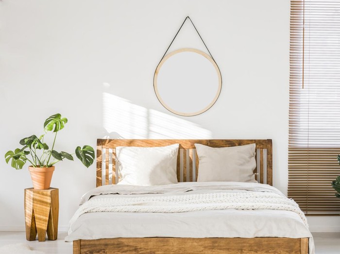 Sun shining on a white wall with a round mirror in a minimalist bedroom interior with natural, wooden furniture and beautiful green plants