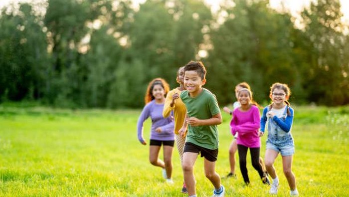 A small group of school aged children are seen running across the grass as they play at recess.  They are each dressed casually and are smiling as they enjoy the fresh air.