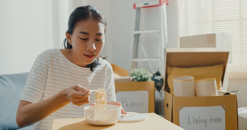 Ilustrasi makan mie instan Young woman moving house sit on couch eating instant noodles after unpacking.